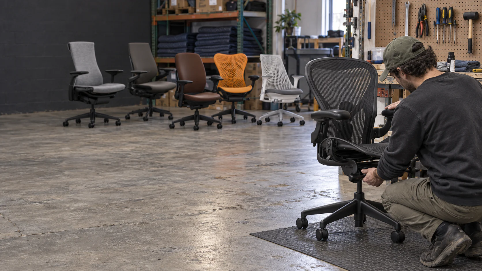 Technician inspecting a black mesh chair with a lineup of classic ergonomic office chairs behind him